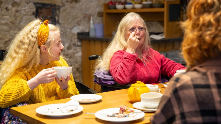 A young girl with long blonde hair in a yellow jumper is sitting at a table holding a mug of hot chocolate. Next to her is an older lady in a pink jumper with long blonde hair and glasses. On the table in front of them both is a yellow teapot and several plates of cakes and scones.